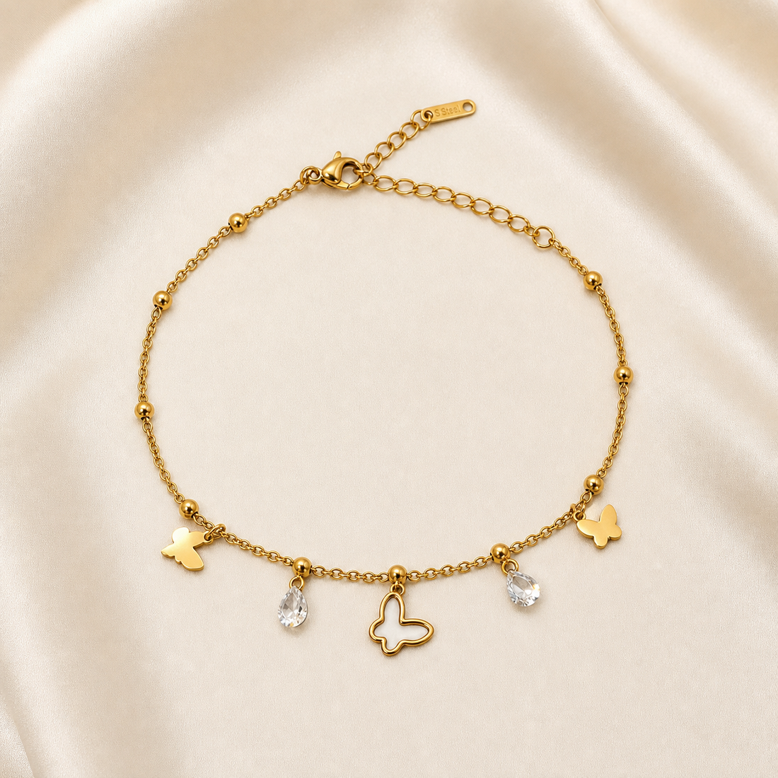 One anklet in gold-tone metal with a polished finish. It is a single-strand fine cable chain of small oval links, punctuated at intervals by small round gold bead stations. The chain run is roughly 25 to 30 times the width of the largest charm; the extender chain is about one quarter the length of the main chain. A working lobster clasp is visible at one end, with an oval-link extender chain terminating in one rectangular engraved metal tag.