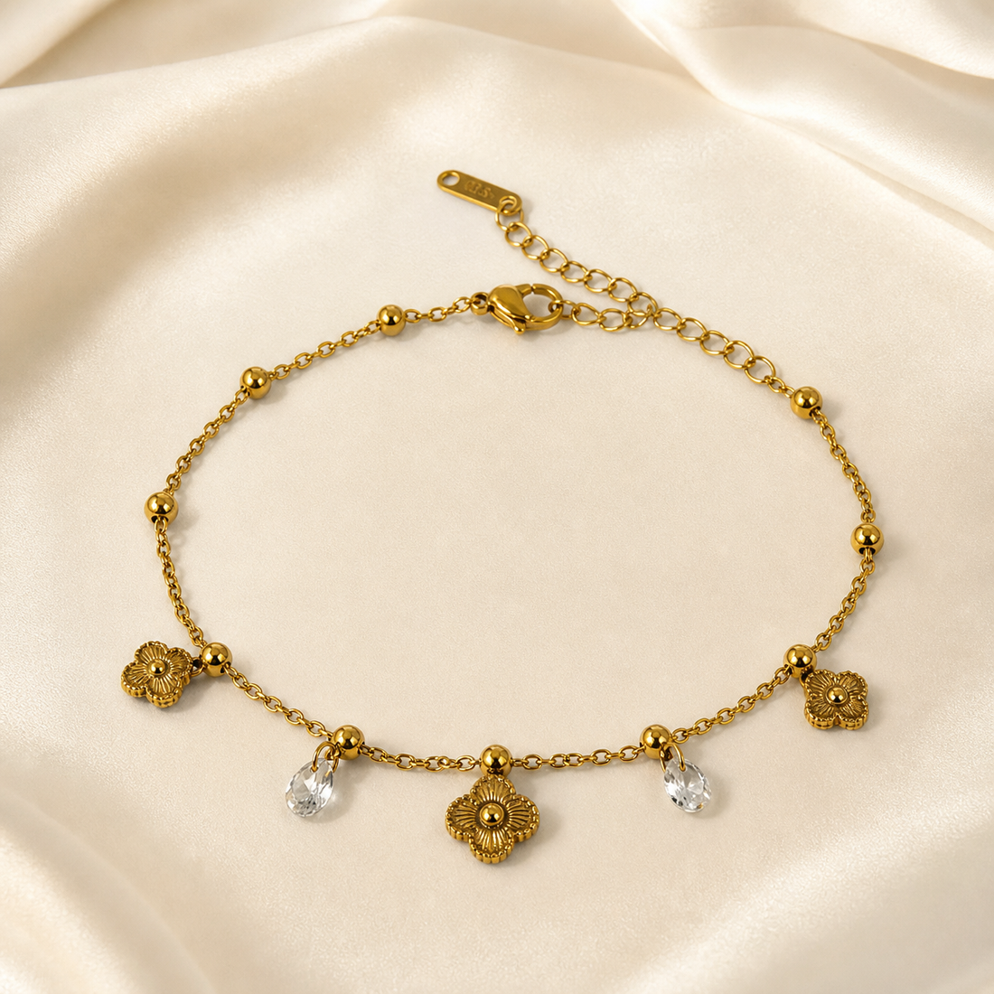 One anklet in yellow-gold-toned metal with a polished finish, built as a single strand of fine-gauge oval-link cable chain. The main chain run measures roughly 20 to 25 times the width of the largest component (the central flower drop), and the extender chain is about one quarter of the main chain length. A working lobster clasp is visible, closing to an oval-link extender chain terminated by a single elongated stamped metal tag.