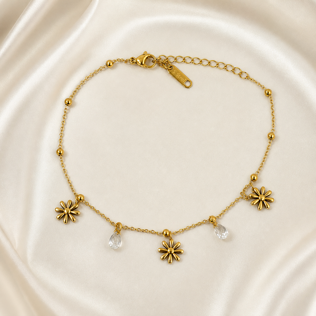 One anklet in yellow-gold-tone polished metal. It is a single strand of fine cable chain with small round bead stations threaded along its length. The chain run measures roughly twenty to twenty-five times the width of the largest component, the flower charm, and the curb-link extender is about one third of the main chain length, ending in a flat elongated stamped terminal tag; a working lobster-claw closure is present and clearly visible.