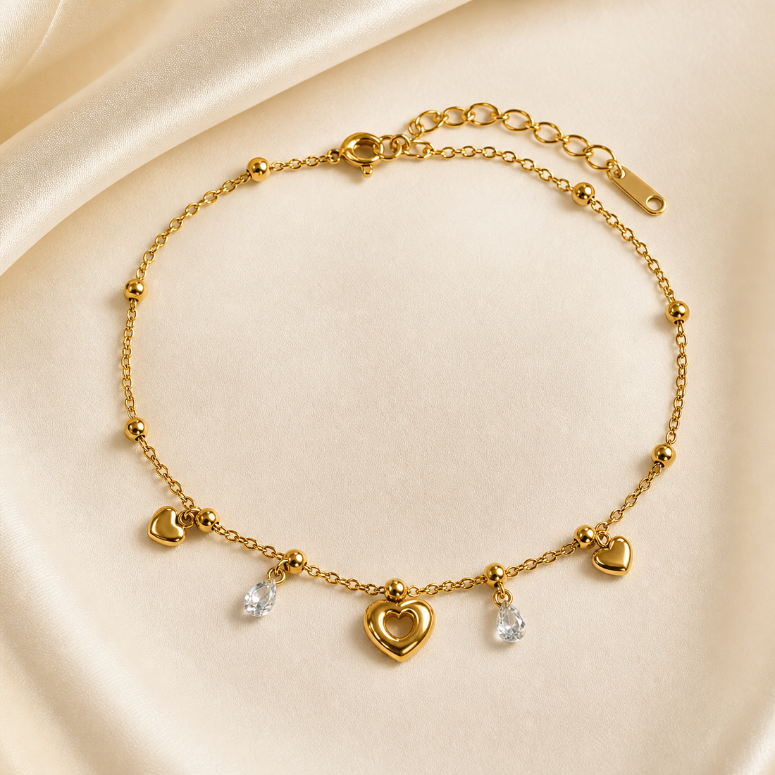 One anklet in gold-coloured metal with a polished finish. It is a single flexible strand of fine cable chain interspersed with small polished round bead stations, with one slightly thicker extension run of larger oval links at the clasp end. The chain run is roughly 25 times the width of the largest component, the central open-heart charm, and the extender is about one sixth of the main chain.