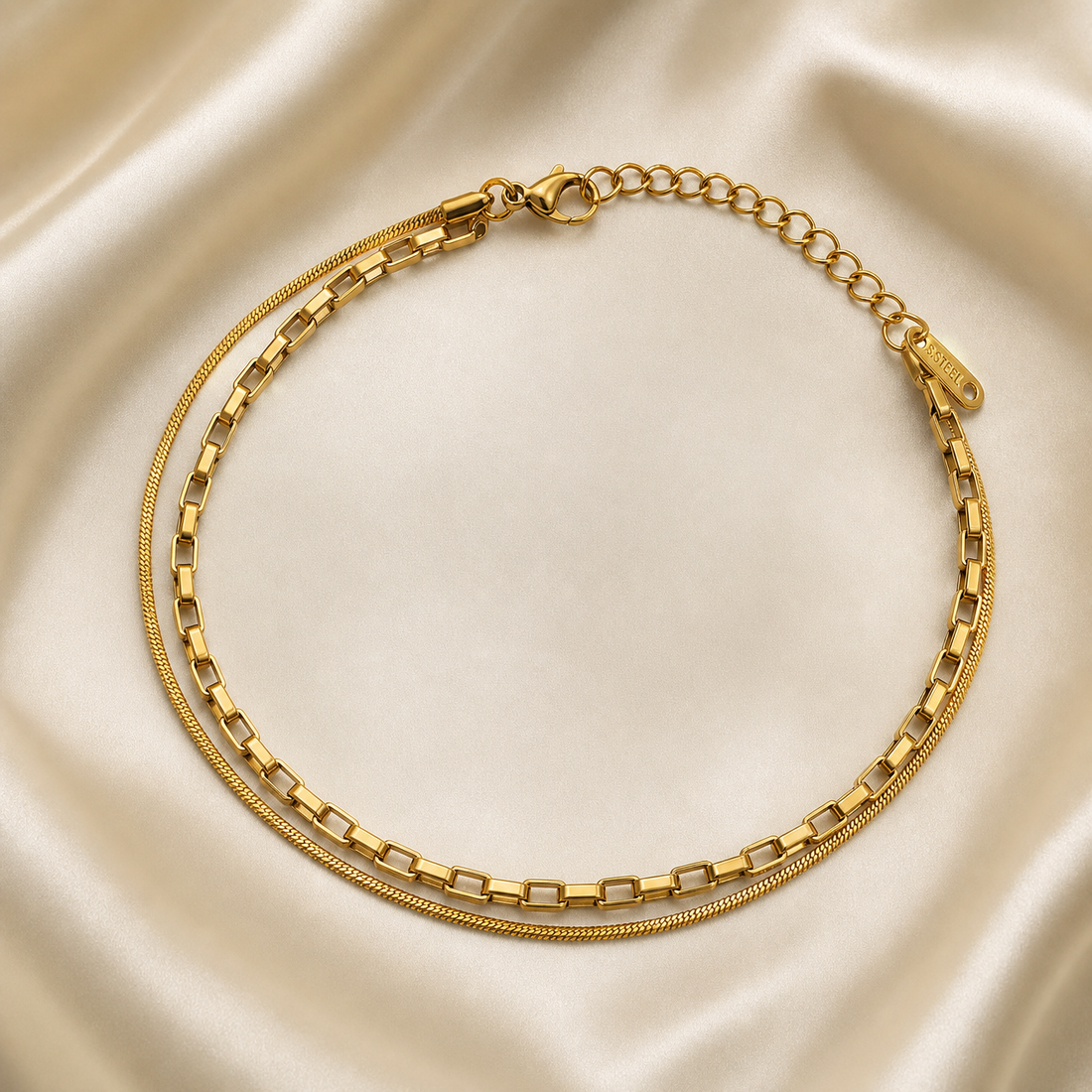 One anklet in gold-toned metal with a bright polished finish. It has two parallel strands joined at shared end fittings: a fine snake chain and a slightly heavier chain built from elongated rectangular box links, many of which are hollow or open-sided, with a small number of links appearing to hold clear rectangular elements that are too small to resolve fully; no charms, drops, bells, coins, tassels, beads or motifs are present, and the construction is uniform along the full length with no separately