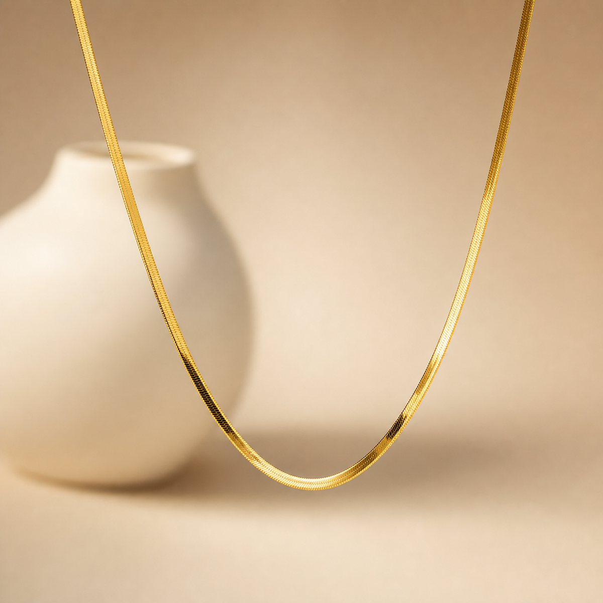 A single necklace of yellow-gold tone metal with a polished finish, built as one continuous flat herringbone chain of fine, tightly interlocking links giving a smooth, ribbon-like band of uniform gauge. The chain has a single strand with no branches, joins or secondary strands, and no pendants, charms, stations, beads or set stones appear anywhere along its length; the surface is plain and continuously decorated only by the herringbone link texture.