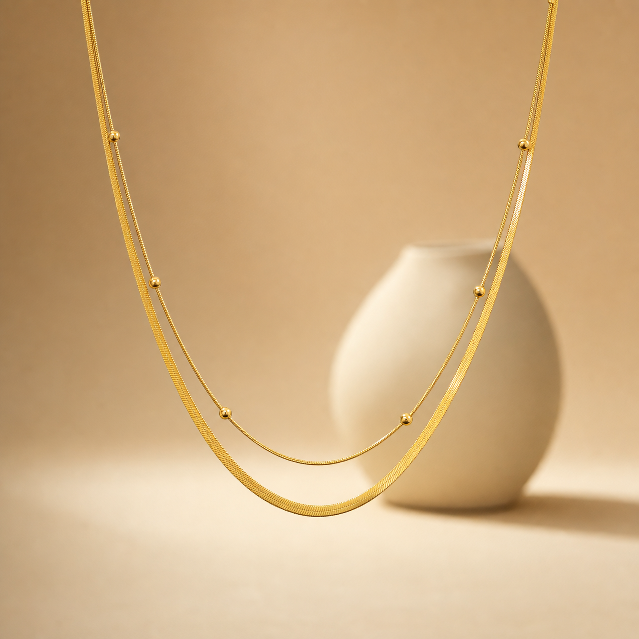 One necklace of yellow-gold-tone metal with a polished finish, built as two joined parallel strands of differing chain type and gauge: a wide flat herringbone chain and a much finer round snake chain, the two sharing common end connections. The full chain run is roughly 60 to 70 times the width of the widest bead component; no extender chain is visible, so no extender fraction can be stated, and no working clasp is resolved in view (the fastening area is partly obscured).