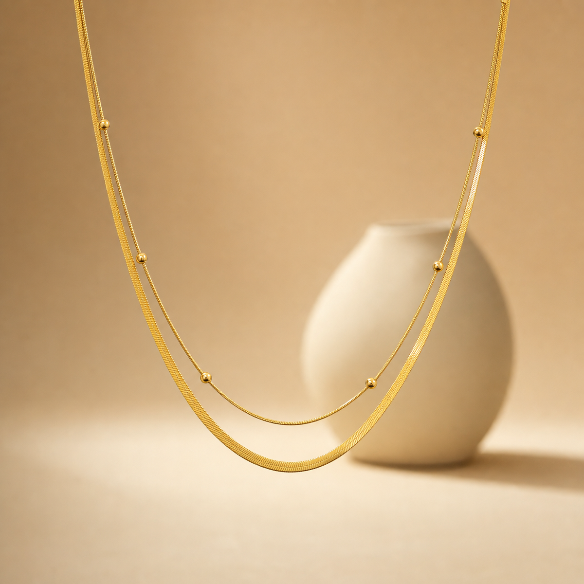One necklace of yellow-gold-tone metal with a polished finish, built as two joined parallel strands of differing chain type and gauge: a wide flat herringbone chain and a much finer round snake chain, the two sharing common end connections. The full chain run is roughly 60 to 70 times the width of the widest bead component; no extender chain is visible, so no extender fraction can be stated, and no working clasp is resolved in view (the fastening area is partly obscured).