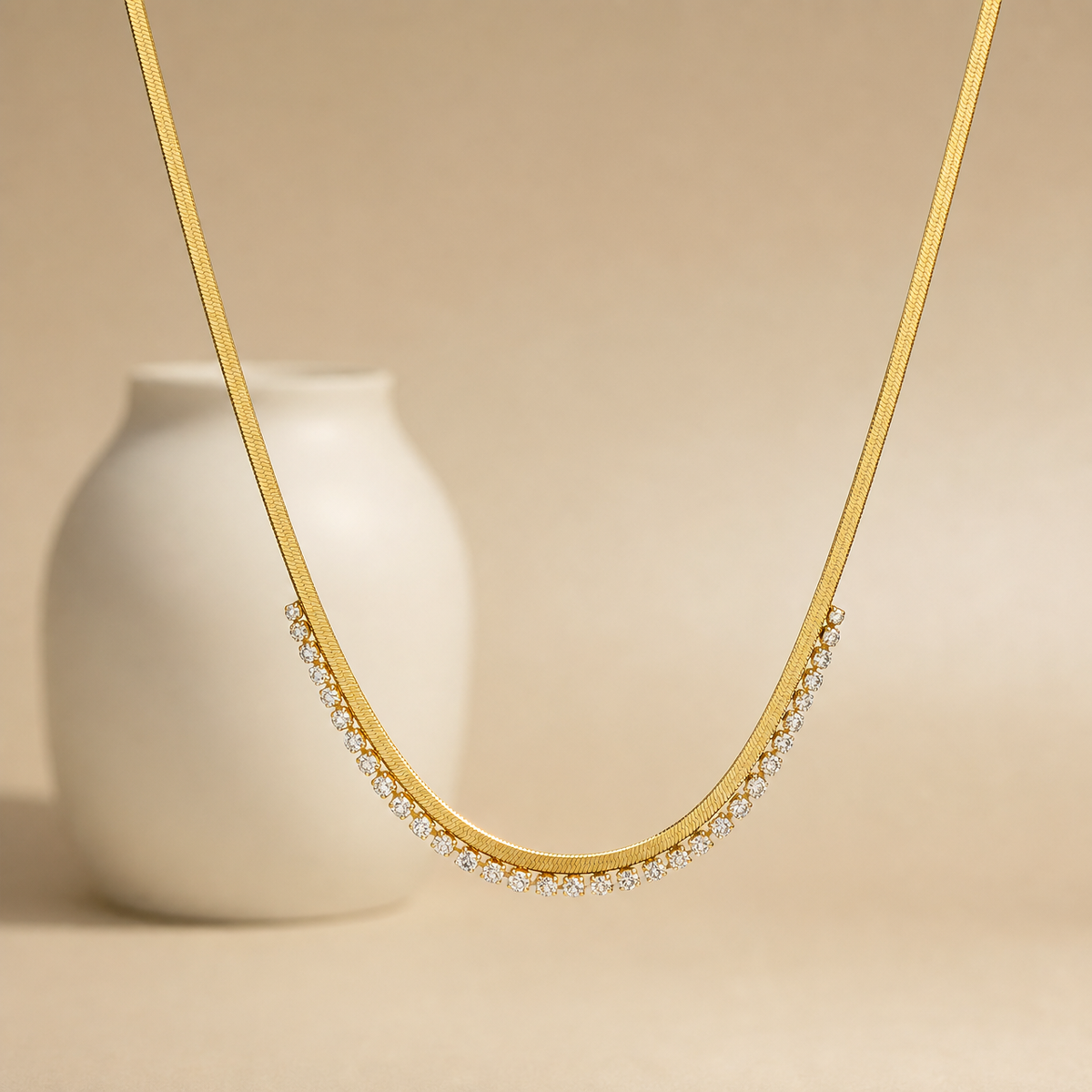 Gold-tone 304 stainless steel herringbone chain necklace with a row of clear crystals set along the front curve, shown laid against a beige backdrop
