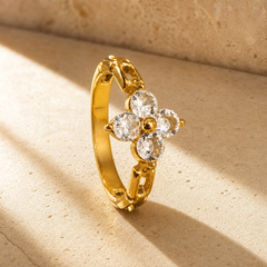 One finger-worn ring in yellow gold with a polished finish. The band is rounded in profile, roughly one-third the width of the head, a closed circle with ornate pierced, bead-tipped shoulders and gallery sections meeting the head. The head is a four-petal flower in raised relief: four round faceted clear stones of equal size, each prong-set around a central solid gold bead, with small gold prong tips extending between the petals. Pierced cut-out openings appear in the shoulder metal.
