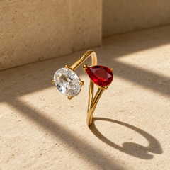 One finger-worn ring in polished yellow gold with a rounded, moderately slender band, roughly one-quarter the width of the head, formed as an open adjustable bypass band whose two ends each carry a stone. The head comprises two prong-set faceted stones facing one another: one oval-cut clear colourless stone and one pear-cut red stone of similar size, each held by four rounded prongs. The clear stone shows visible internal inclusions.