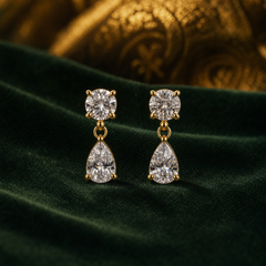 Two earrings of one design, in polished yellow-gold-tone metal. Each fits the ear with a post stud, the backs not visible. From the stud downward: a round colourless faceted clear stone in a four-prong setting, then a small metal ball and open jump ring, then a larger pear-shaped colourless faceted clear stone in a prong setting with V-tip, dangling freely from the ring. The drop is roughly two and a half times the widest part, the pear stone.