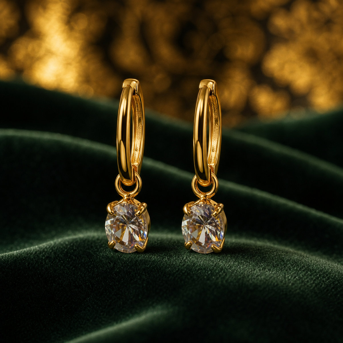 Two ear-worn earrings, one design in gold-toned polished metal. Each has a small huggie-hinge hoop fitting, a narrow round band of fine gauge. From the base of each hoop hangs, via a small jump ring, a single clear round faceted stone held in a gold-toned multi-prong basket setting. Each earring has two sections — hoop and stone dangle — hinged at the jump ring. Overall drop is roughly two-and-a-half times the widest part, the stone.