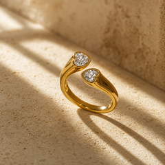 Two finger-worn rings of matching design in yellow-gold-toned metal with a polished finish. Each has a rounded band, roughly half the width of its head, forming a closed circle. Each head is a single heart-shaped bezel holding one heart-shaped faceted clear stone in a bezel setting, the stone's face raised slightly above the metal rim; the two stones are similar in size, one marginally larger. The shoulders taper smoothly into the bezel with no pavé or accent stones.