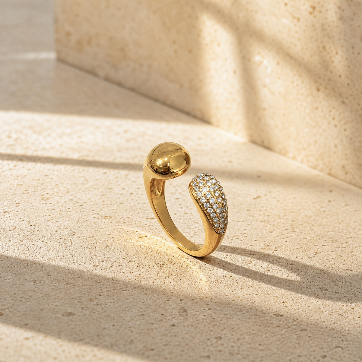 One finger-worn ring in yellow-gold-toned metal with a polished finish. It is an adjustable open band of rounded profile, roughly a third the width of the larger terminal, its two ends facing each other across a gap. One terminal is a smooth, highly reflective solid-metal dome; the other is a teardrop-shaped head densely covered in small round faceted clear stones in a continuous pavé field, stones too numerous and small to count individually.