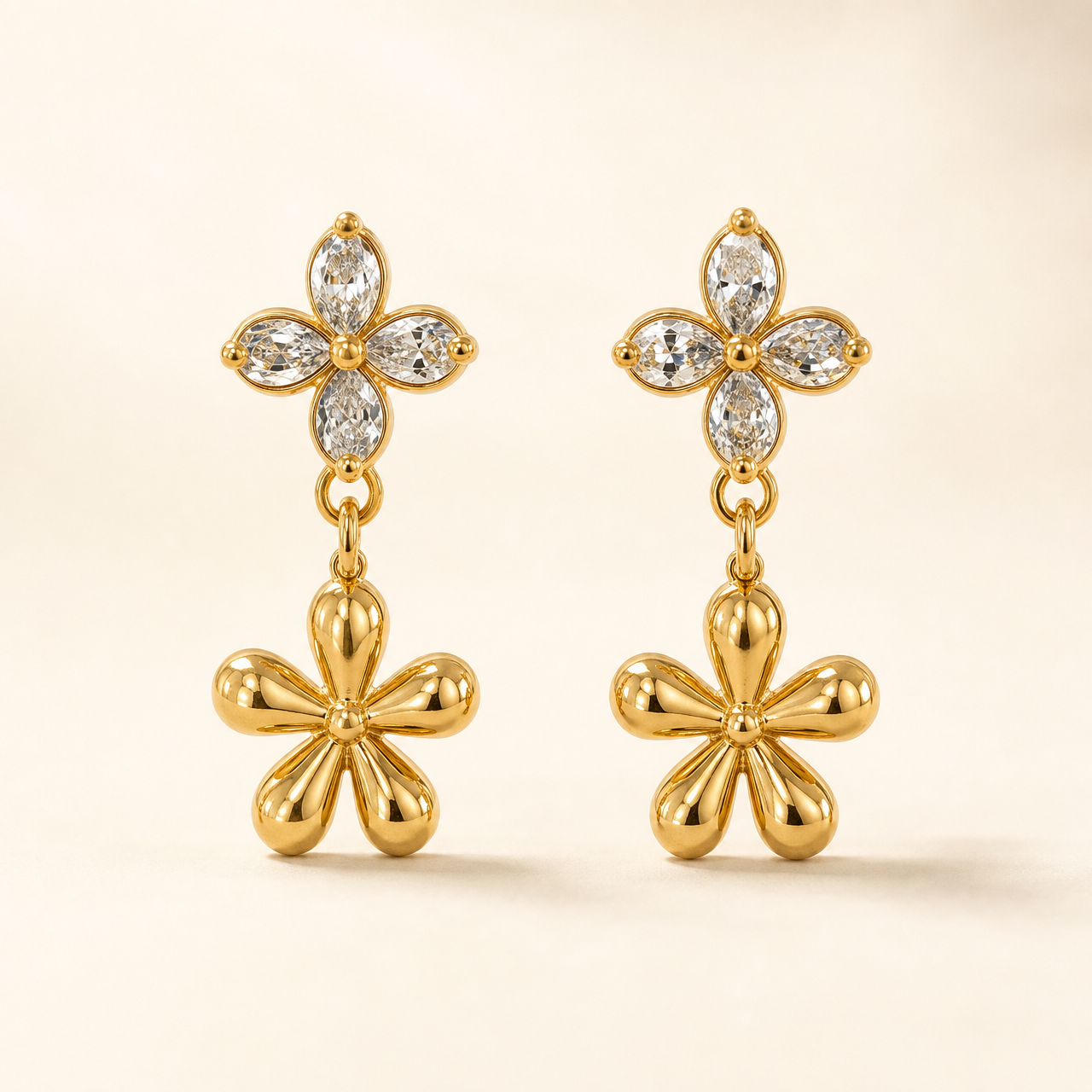 Two ear-worn earrings of one design in polished yellow-gold-tone metal. Each carries a post fitting with a friction back; the back is partly obscured, post gauge slim and length standard. Total drop is roughly twice the width of the widest motif. Two articulated sections: an upper four-petal flower stud of clear pear-cut faceted stones prong-set around a small gold bead centre, and below it a larger solid polished-gold five-petal flower dangling freely from a small link.