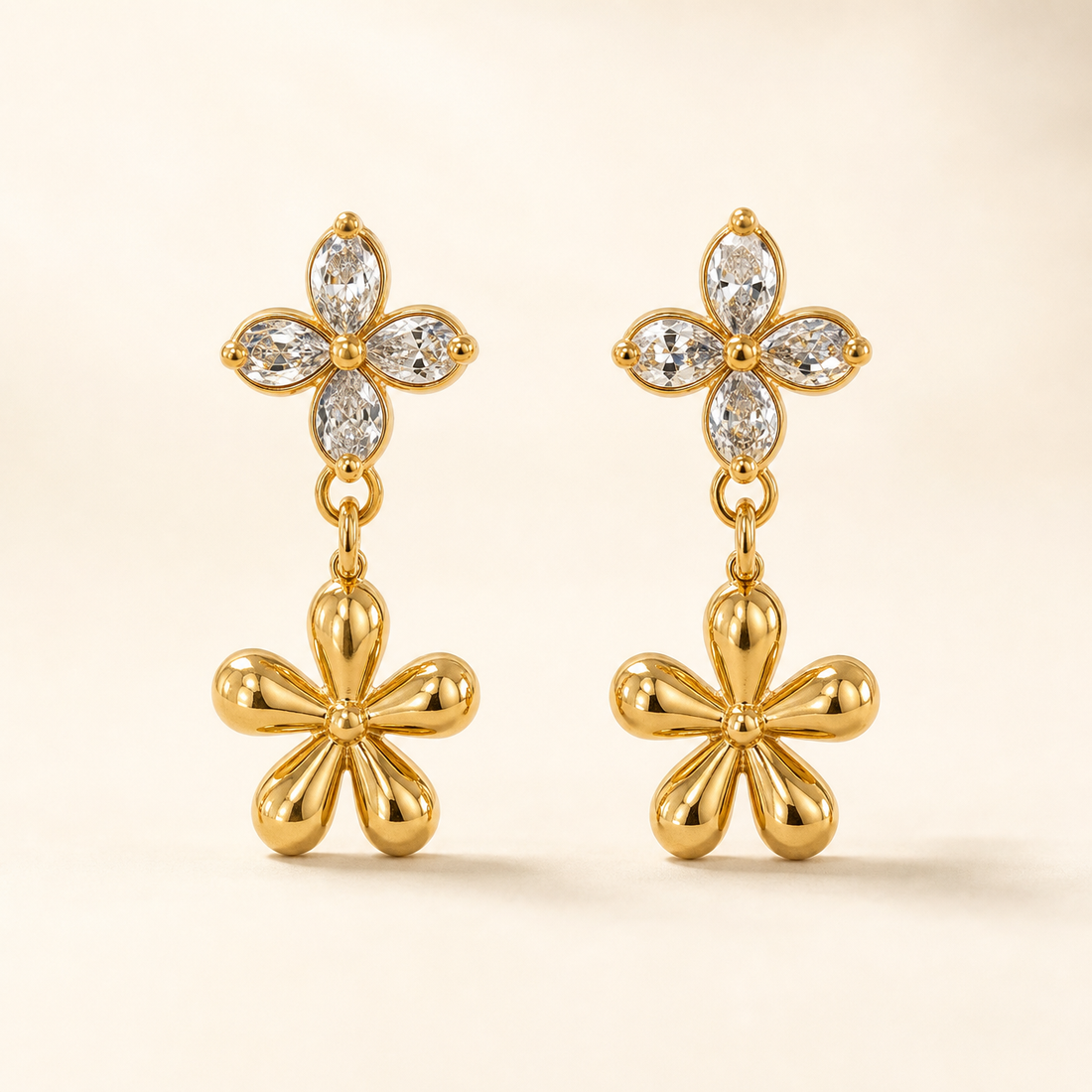 Two ear-worn earrings of one design in polished yellow-gold-tone metal. Each carries a post fitting with a friction back; the back is partly obscured, post gauge slim and length standard. Total drop is roughly twice the width of the widest motif. Two articulated sections: an upper four-petal flower stud of clear pear-cut faceted stones prong-set around a small gold bead centre, and below it a larger solid polished-gold five-petal flower dangling freely from a small link.