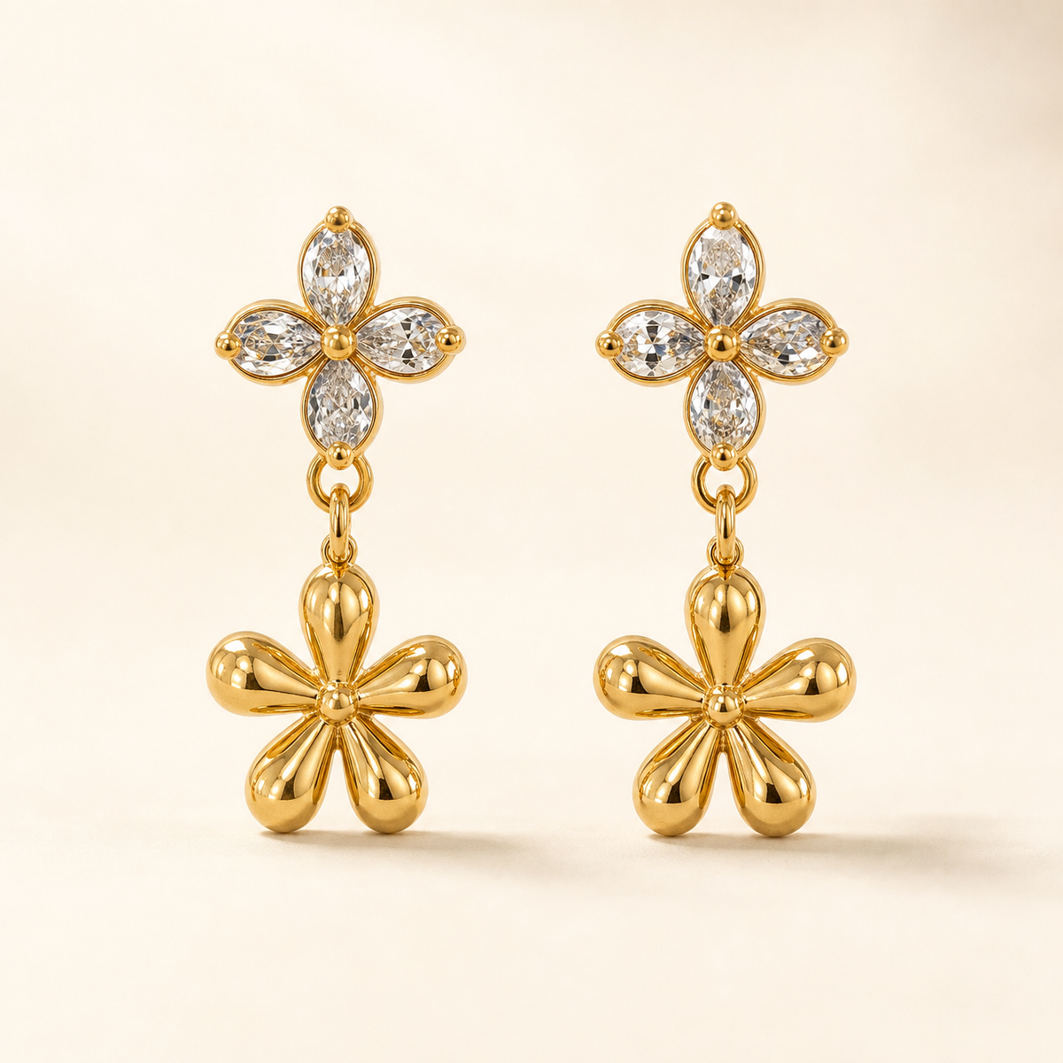 Two ear-worn earrings of one design in polished yellow-gold-tone metal. Each carries a post fitting with a friction back; the back is partly obscured, post gauge slim and length standard. Total drop is roughly twice the width of the widest motif. Two articulated sections: an upper four-petal flower stud of clear pear-cut faceted stones prong-set around a small gold bead centre, and below it a larger solid polished-gold five-petal flower dangling freely from a small link.
