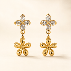 Two ear-worn earrings of one design in polished yellow-gold-tone metal. Each carries a post fitting with a friction back; the back is partly obscured, post gauge slim and length standard. Total drop is roughly twice the width of the widest motif. Two articulated sections: an upper four-petal flower stud of clear pear-cut faceted stones prong-set around a small gold bead centre, and below it a larger solid polished-gold five-petal flower dangling freely from a small link.