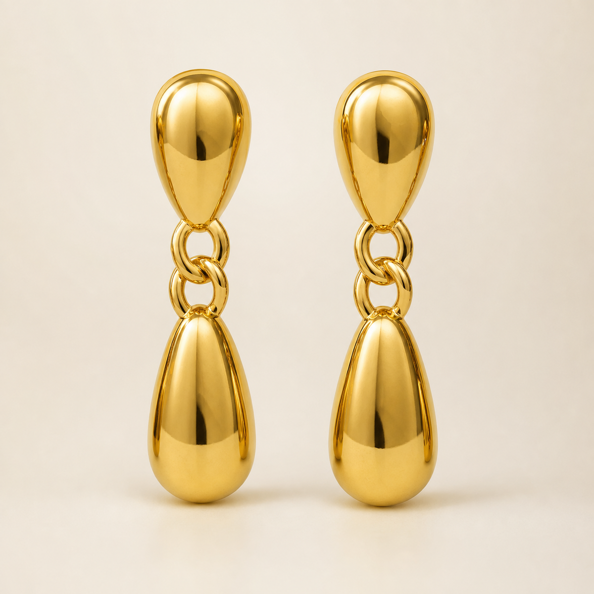 Two ear-worn earrings of one design in yellow-gold-tone metal with a high-polish mirror finish. Each is a drop earring on a post fitting (the posts and backs are partly obscured) emerging from a domed, hollow teardrop stud. From the stud hangs a short articulated chain of two interlocking oval links, ending in a larger solid-metal teardrop dangle, so each earring has three sections hinging at two joints. The total drop is roughly three times the widest teardrop width.