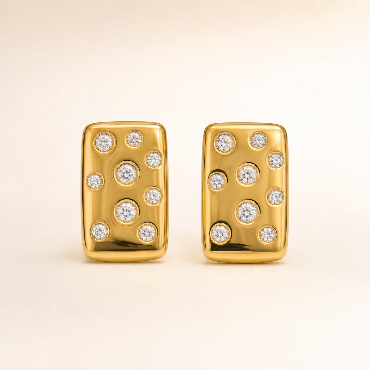 Two ear-worn stud earrings of one design, in yellow-gold-tone metal with a polished finish. Each earring carries a post-and-back fitting (post and clutch partly obscured by the display mount). Each is a single rigid section, a slightly domed rectangular plaque with rounded corners, its face scattered with round clear faceted stones in bezel-style flush settings of varying small sizes, one noticeably larger stone near the middle of each plaque.