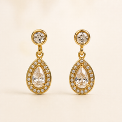 Two ear-worn earrings of one design in yellow-gold-toned metal with a polished finish. Each has a post fitting (back not visible, likely a standard push post), the post topped by a small round clear faceted stone in a smooth bezel. From the stud a short jump-ring link articulates to a teardrop-shaped drop section, giving two hinged sections; overall drop is roughly two and a half times the widest point (the teardrop).