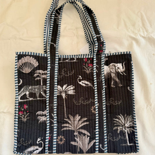 Black quilted canvas tote bag with a jungle print of leopard, elephant, palms and flamingos, and striped blue-and-white trim, lying flat