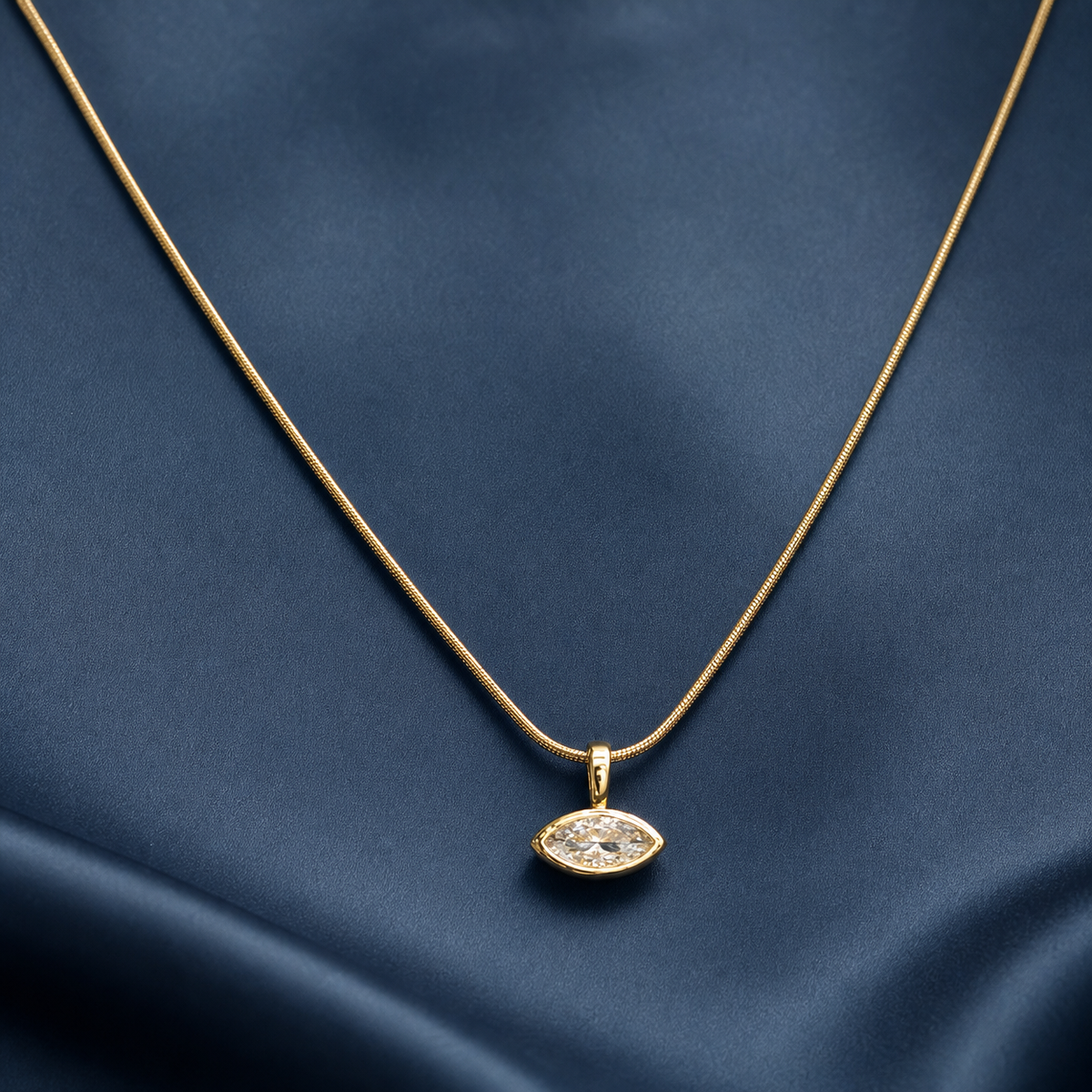 Gold-tone 316L stainless steel necklace with a thin snake chain and a marquise crystal pendant set in a bezel, laid on dark blue fabric
