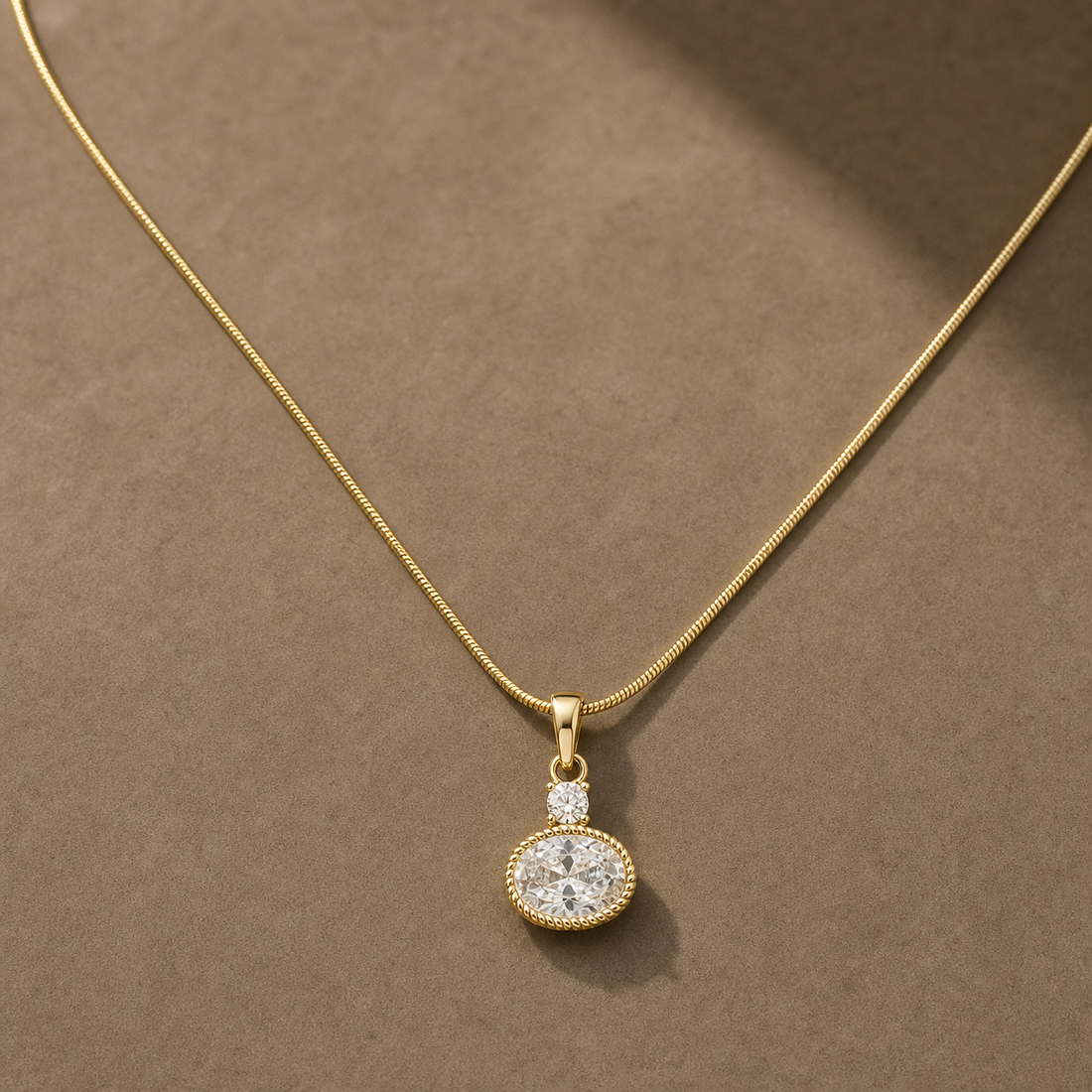 Gold-tone 316L stainless steel snake chain necklace with an oval crystal pendant and a small round crystal above it, laid flat on a neutral surface