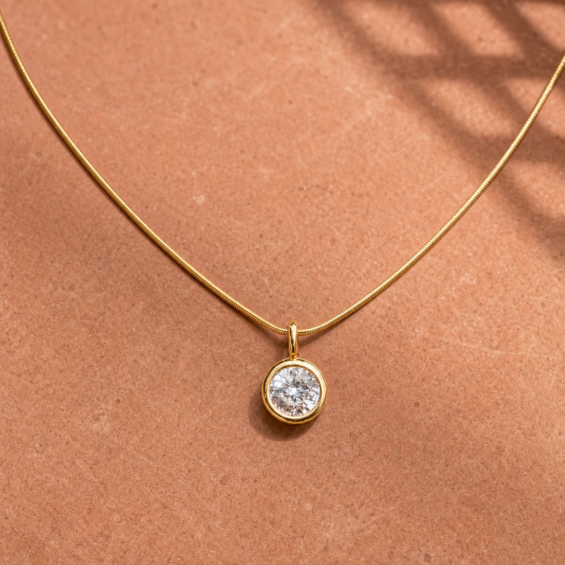 Gold-tone 316L stainless steel necklace with a fine snake chain and a round clear stone pendant in a bezel setting, laid flat on a terracotta surface