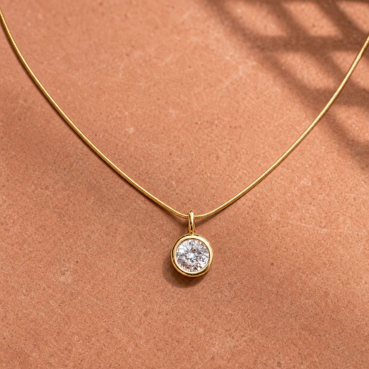 Gold-tone 316L stainless steel necklace with a fine snake chain and a round clear stone pendant in a bezel setting, laid flat on a terracotta surface