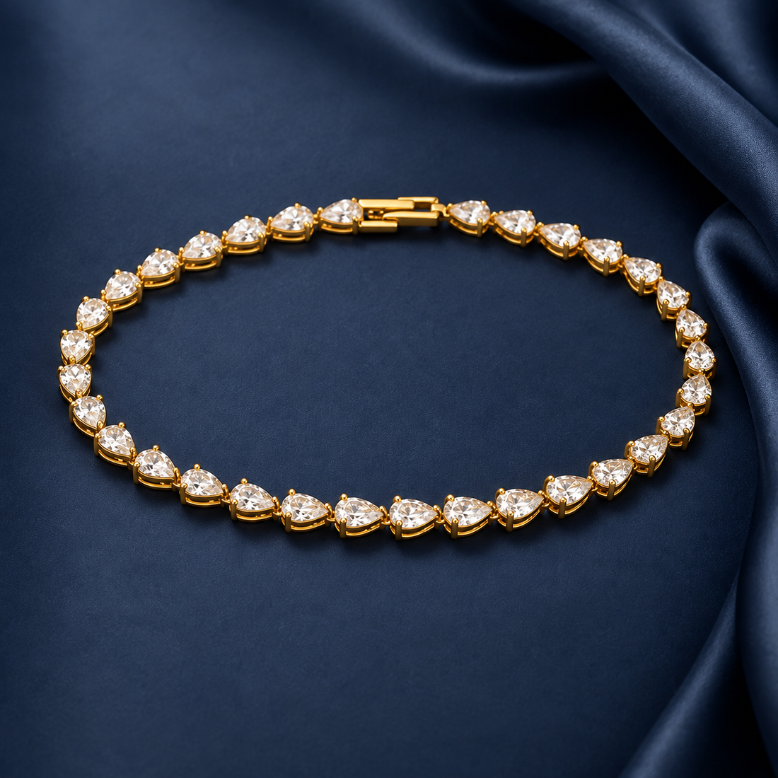 Gold-tone brass tennis bracelet with pear-cut clear stones in prong settings and a box clasp, laid flat on dark blue fabric