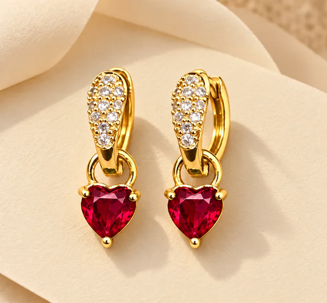 Gold-tone 316L stainless steel huggie earrings with pavé crystals on the hoop and a red heart-cut stone drop, a pair shown flat on a cream surface