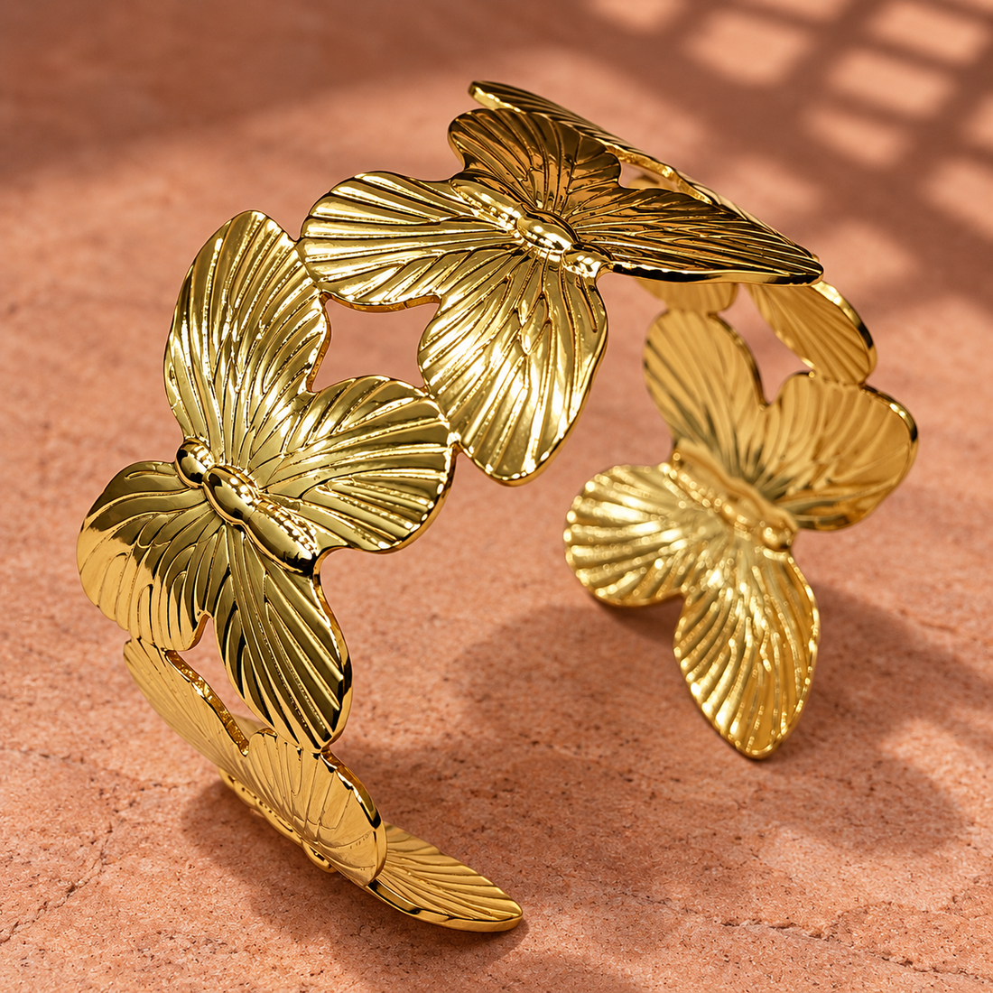 Gold-tone 304 stainless steel cuff bracelet with a row of embossed butterfly motifs and open ends, shown on a terracotta surface