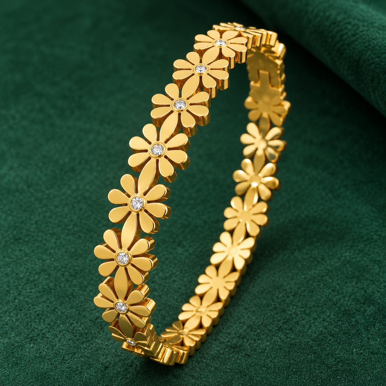 Gold-tone 304 stainless steel kada bracelet with a row of daisy motifs and a clear stone at each flower centre, lying on a green surface