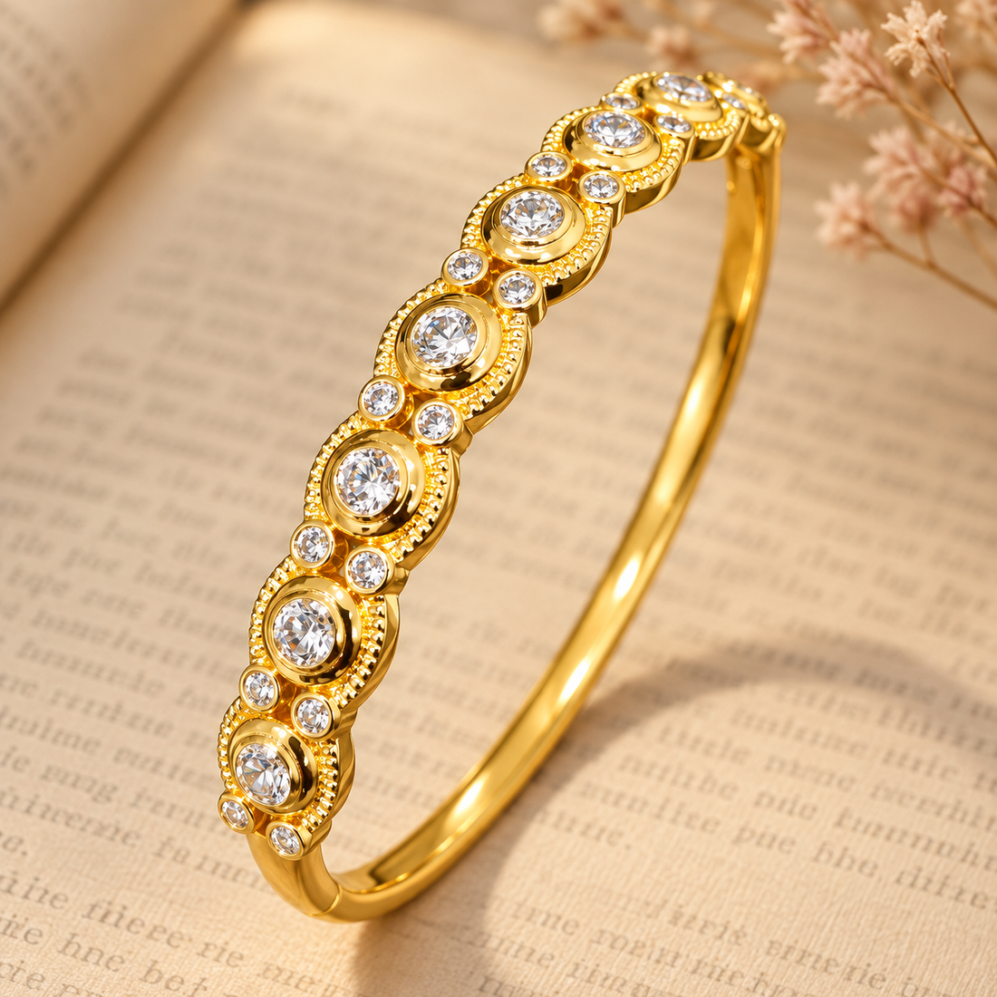 Gold-tone 316L stainless steel kada bracelet with a row of round clear stones in milgrain-edged bezel settings along the front, shown lying on an open book
