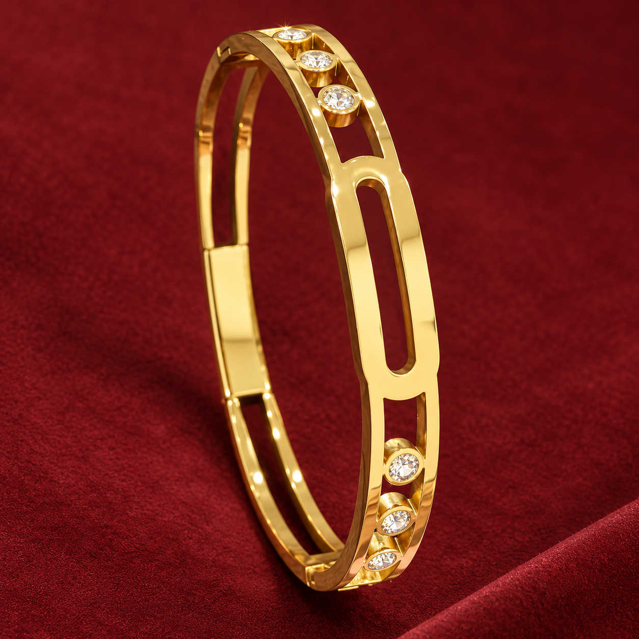 Gold-tone 304 stainless steel bangle with an open oval link design and crystal-set bezels, shown lying on a deep red fabric surface