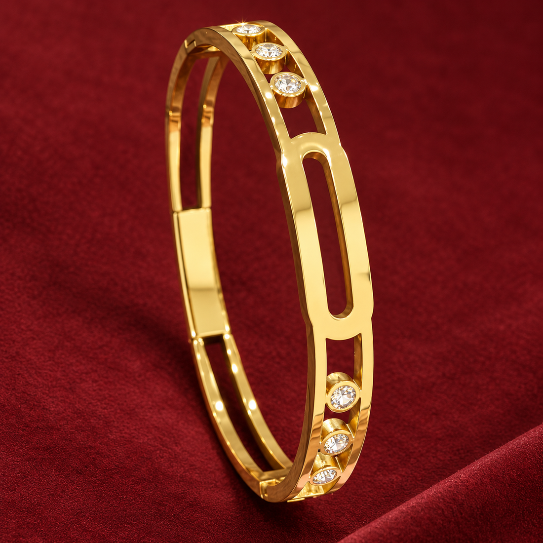Gold-tone 304 stainless steel bangle with an open oval link design and crystal-set bezels, shown lying on a deep red fabric surface