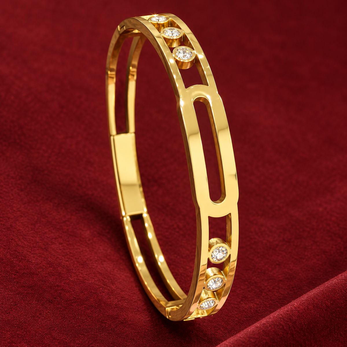 Gold-tone 304 stainless steel bangle with an open oval link design and crystal-set bezels, shown lying on a deep red fabric surface