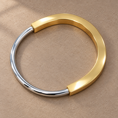 Two-tone 316L stainless steel kada bracelet with a gold-tone wide band and a silver-tone round wire section, lying flat on a beige surface, top view