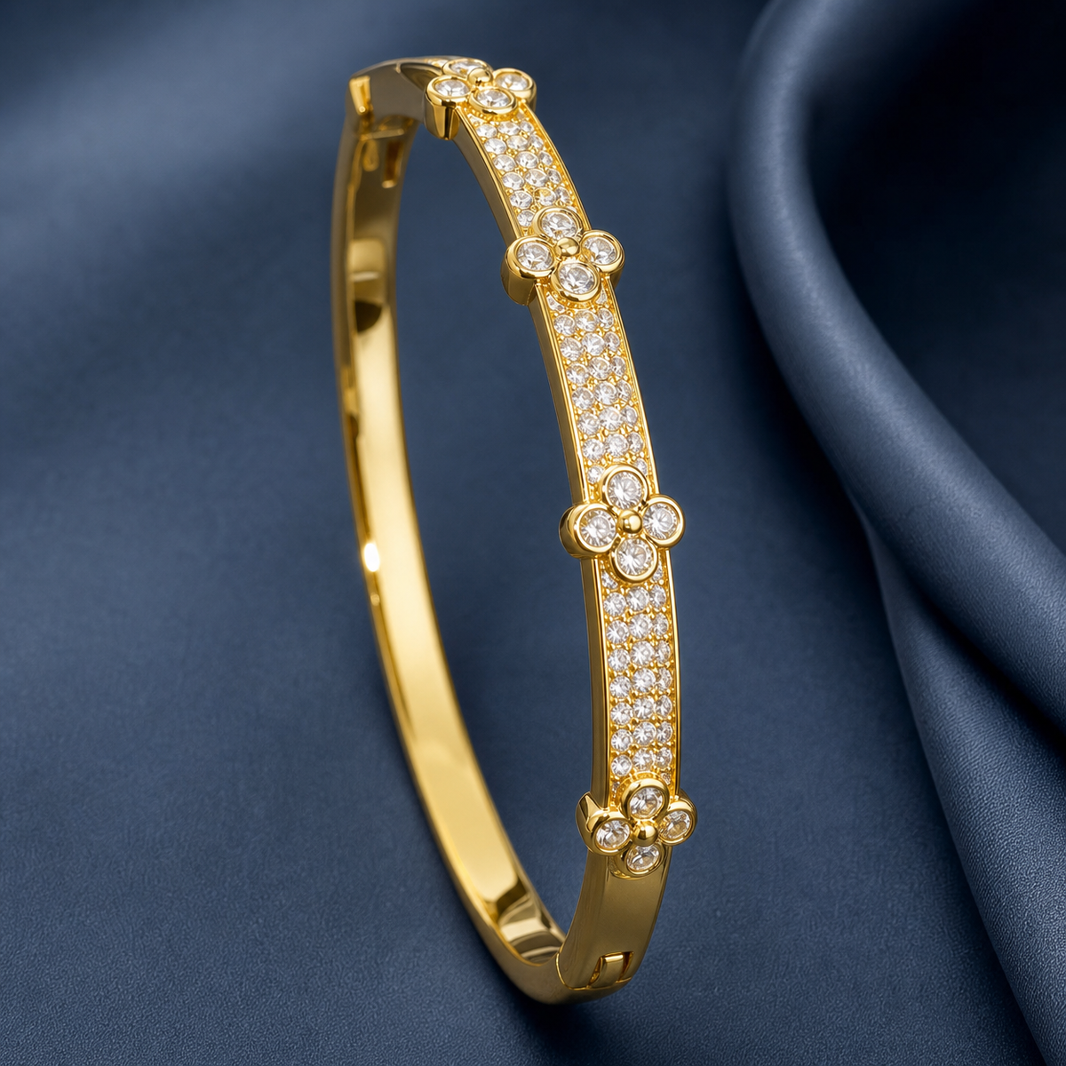 Gold-tone 316L stainless steel kada bracelet with pavé crystal bands and clover-shaped crystal motifs on a hinged bangle, shown on a dark blue fabric