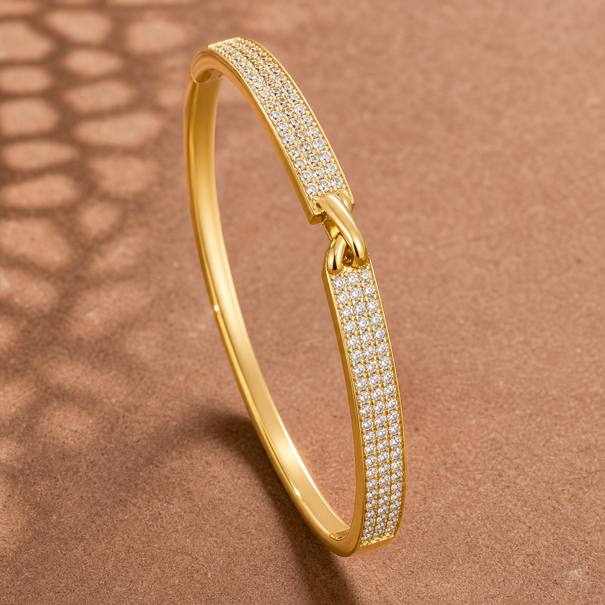 Gold-tone 316L stainless steel kada bracelet with a band set with clear crystals on the top and sides and a knot-style clasp, shown on a plain surface