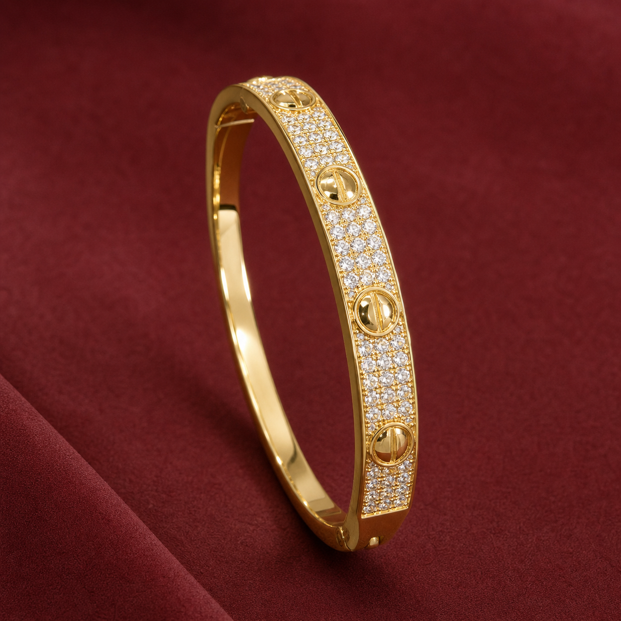 Gold-tone 316L stainless steel bangle bracelet with pavé clear stones across the front and four screw-head motifs, on a red fabric surface