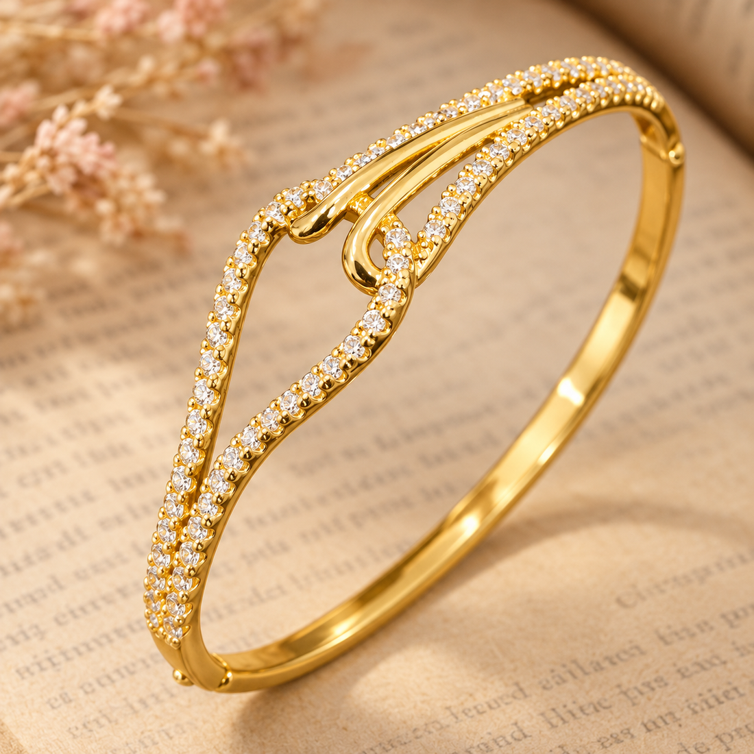 Gold-tone 316L stainless steel kada bracelet with crossed open bands set with clear crystals and a hinged clasp, shown lying on an open book