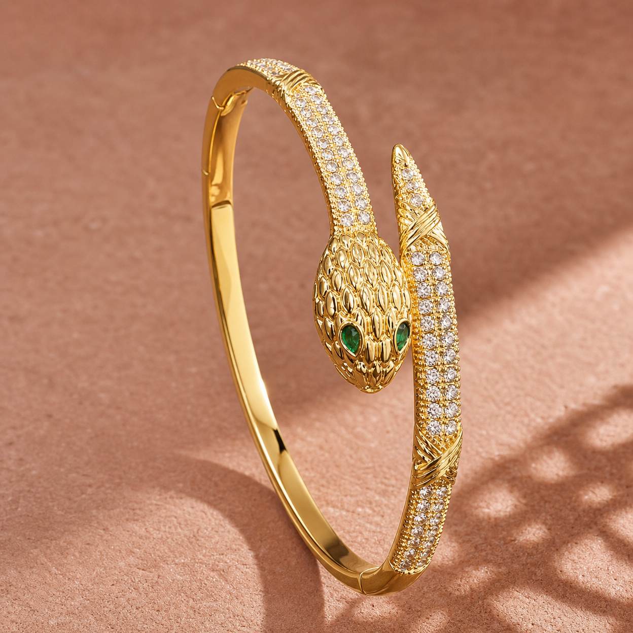Gold-tone 316L stainless steel snake bangle with a scaled serpent head, green stone eyes and a pavé crystal band, on a neutral background
