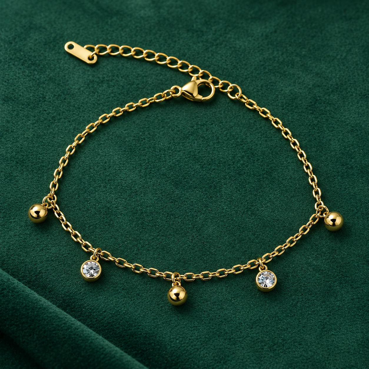 Gold-tone 304 stainless steel chain bracelet with five dangling charms alternating plain ball and clear crystal-set bezel, flat lay on green velvet
