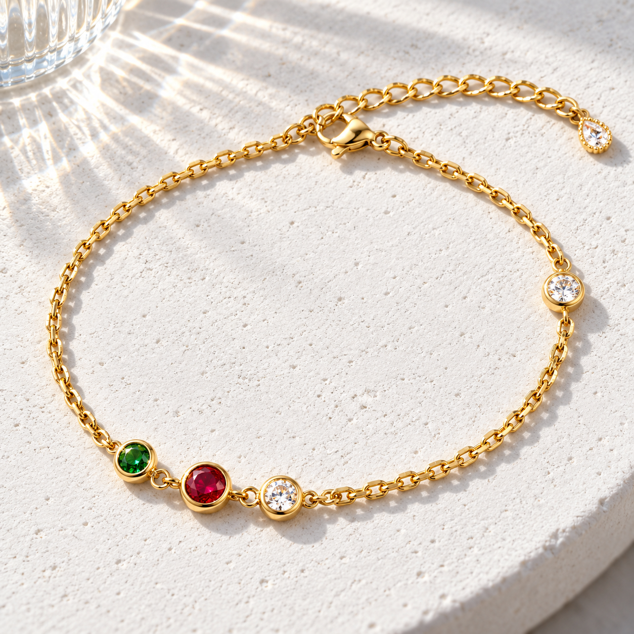Gold-tone 316L stainless steel chain bracelet with bezel-set green, red and clear stones spaced along the links, displayed flat on a stone surface