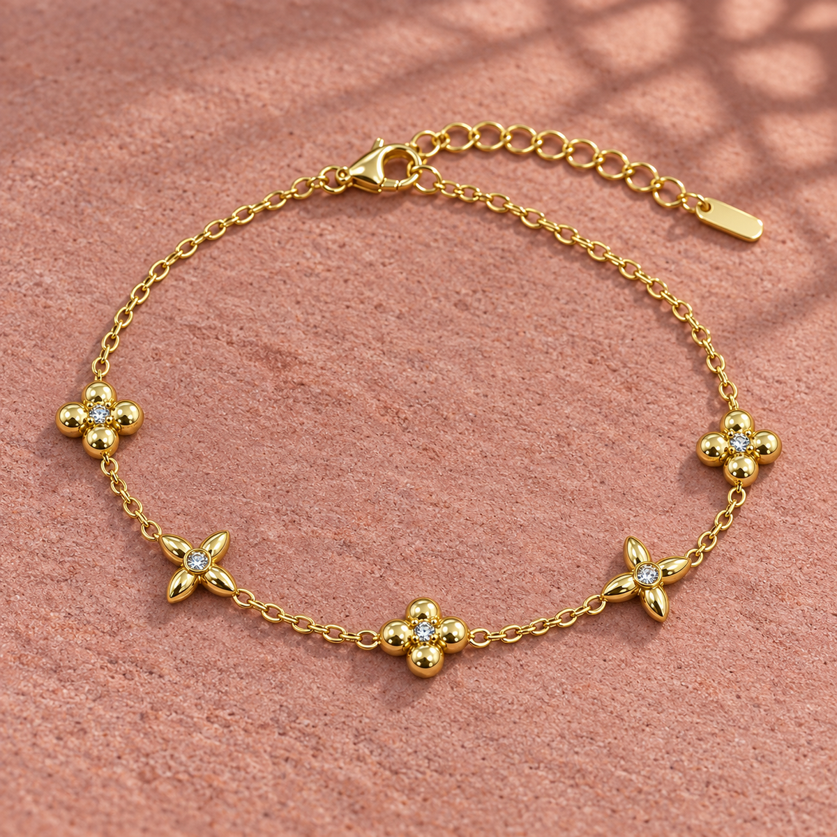 Gold-tone 316L stainless steel chain bracelet with five flower motifs set with small crystals, lying flat on a pink surface