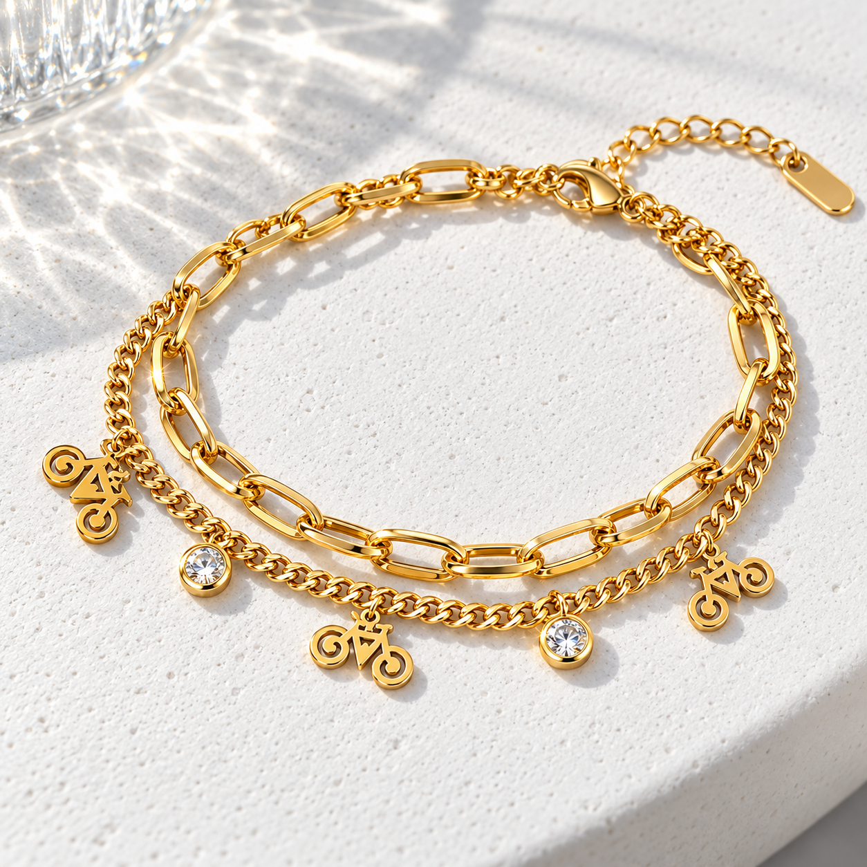 Gold-tone 316L stainless steel double-strand bracelet with paperclip and curb chains, bicycle charms and clear stone drops, lying flat on a white surface