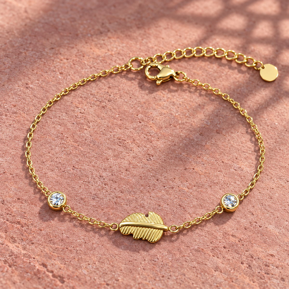 Gold-tone 316L stainless steel chain bracelet with a textured feather charm and two round clear stones, laid flat on a terracotta surface
