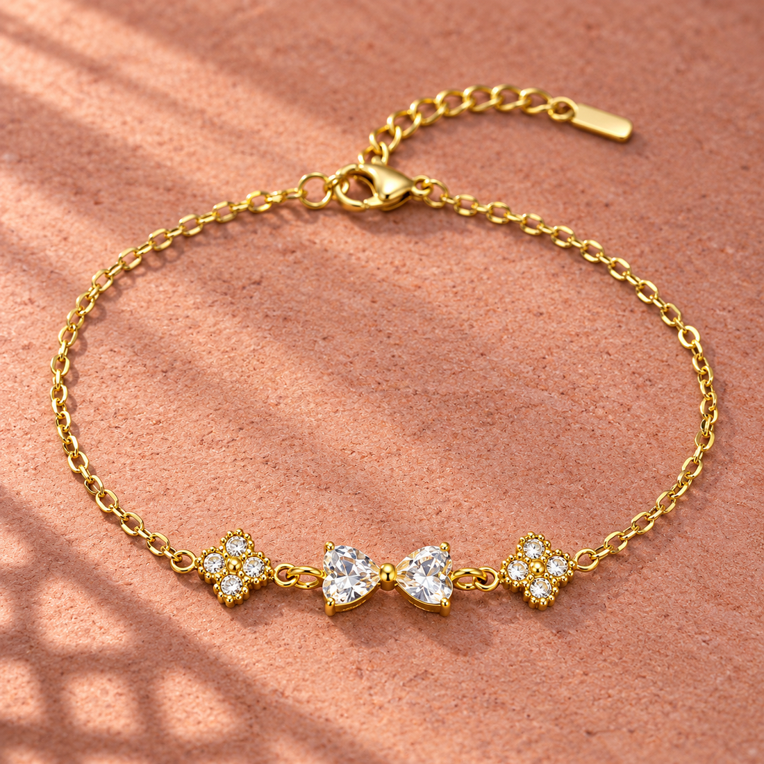 Gold-tone 304 stainless steel chain bracelet with two heart-cut clear stones forming a bow and two four-stone clusters, lying flat on a sandy surface