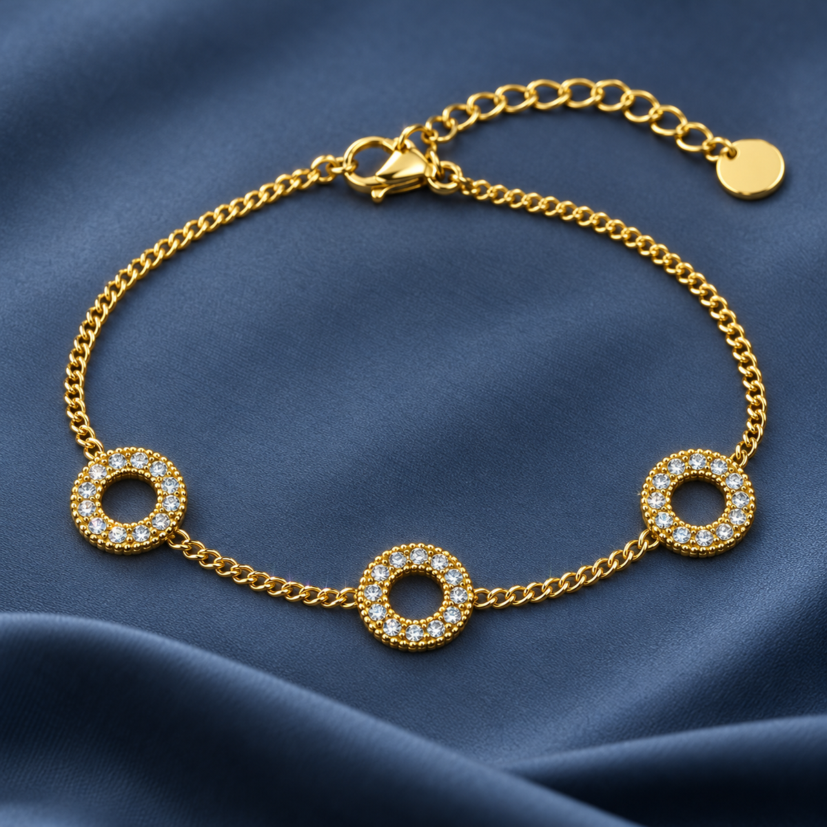 Gold-tone 316L stainless steel chain bracelet with three open circle motifs set with clear stones and a lobster clasp, shown laid flat on blue fabric