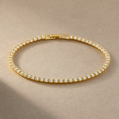 Gold-tone brass tennis bracelet with a row of clear stones in claw settings and a box clasp, laid flat on a beige surface