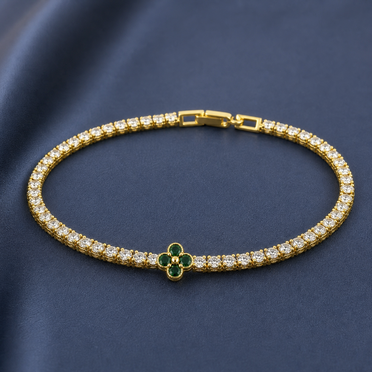 Gold-tone brass tennis bracelet with clear stones and a four-petal clover set with green stones, lying flat on a navy background
